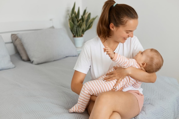 Portrait of happy satisfied attractive woman wearing white t shirt and shorts sitting on bed with her baby daughter, mother playing with her kid, expressing love and gentle.