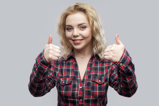 Photo portrait of happy satisfied beautiful blonde young woman in casual red checkered shirt standing and looking at camera with toothy smile and thumbs up. studio shot, isolated on grey background.