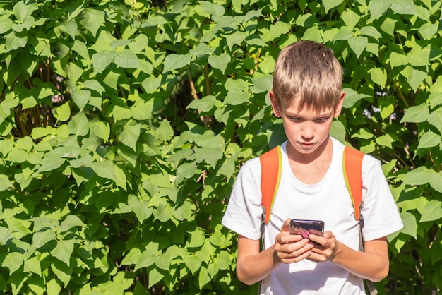 Portrait of a happy schoolboy with a backpack using a smartphone writing online messages on the street