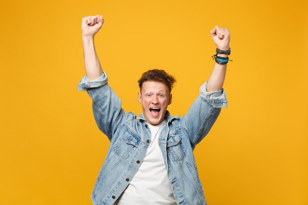 Portrait of happy screaming young man in denim casual clothes clenching fists like winner isolated on yellow orange background in studio. People sincere emotions lifestyle concept. Mock up copy space.