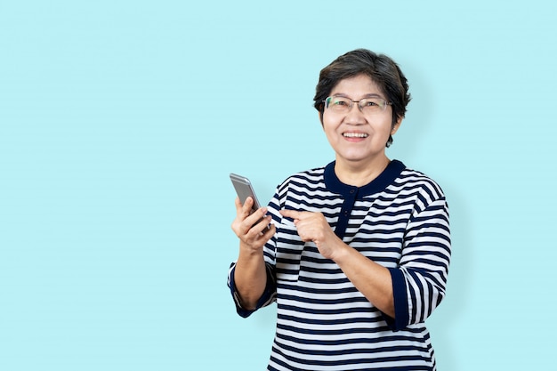 Portrait of happy senior asian woman holding or using smartphone and looking at camera on isolated background feeling positive enjoy and satisfaction. Older female lifestyle concept  blue background.