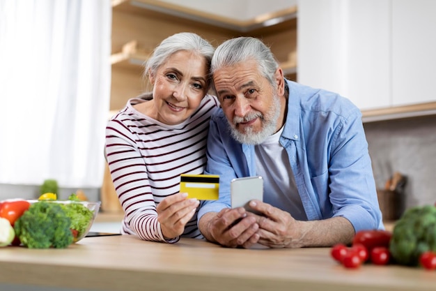 Portrait Of Happy Senior Spouses With Smartphone And Credit Card In Kitchen