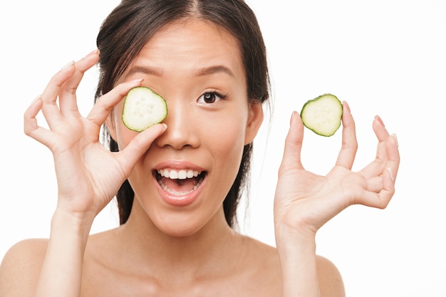 Portrait of a happy shocked excited young beautiful asian woman posing isolated over white wall holding cucumber