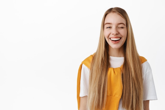 Portrait of happy smiling blond woman with straight long natural blond hair, winking and laughing carefree, standing in t-shirt against white wall