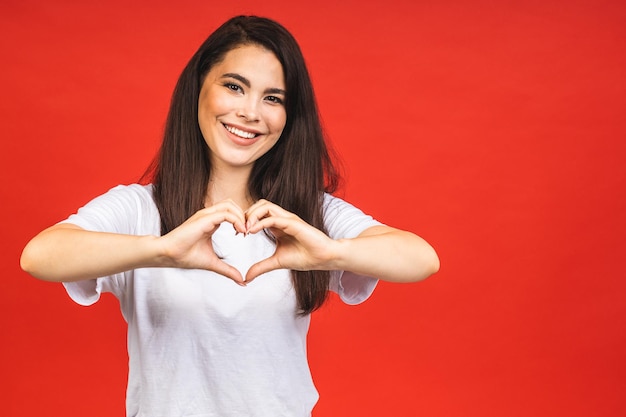 Portrait of happy smiling brunette girl in casual isolated over red background Heart sign with hands