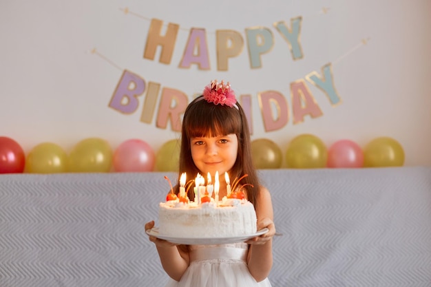 Portrait of happy smiling little girl standing holding cake with candles, celebrating birthday, being ready to make wish and blowing, posing indoor with festive inscription and balloons on background