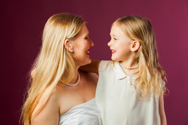 Portrait of happy smiling mother and daughter both with long loose blond hair wearing white clothes and red lipstick Daughter looks like mother Studio with purple background