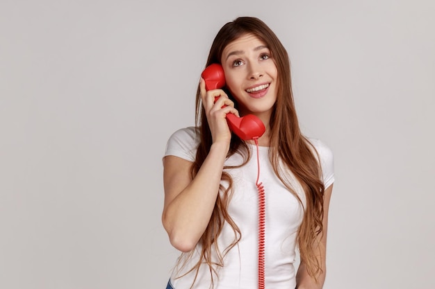 Portrait of happy smiling woman holding red retro phone in hands looking away with dreamy facial expression wearing white Tshirt Indoor studio shot isolated on gray background