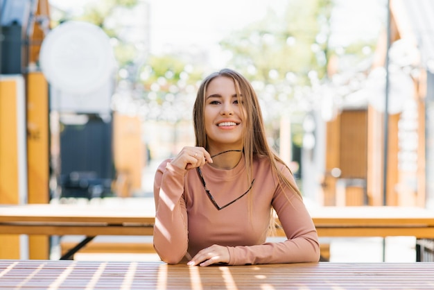 Portrait of a happy smiling woman holding sunglasses in the city in summer in a cafe and waiting f