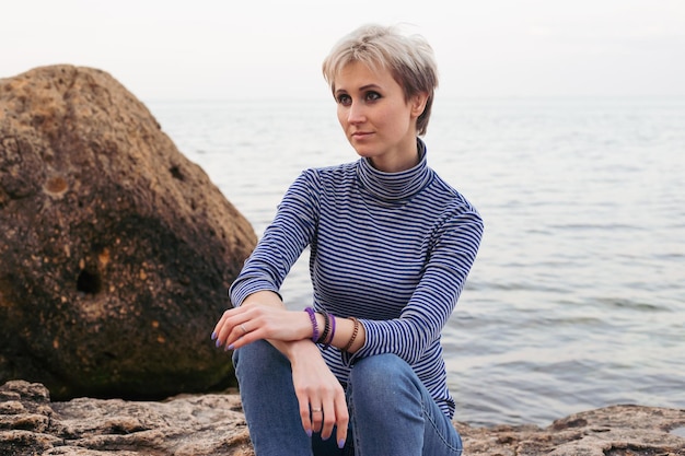 Portrait of happy smiling woman sitting on the rock near sea in autumn or summer time