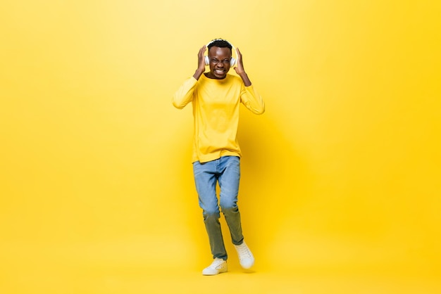 Portrait of happy smiling young African man listening to music from headphones and dancing in yellow studio isolated background