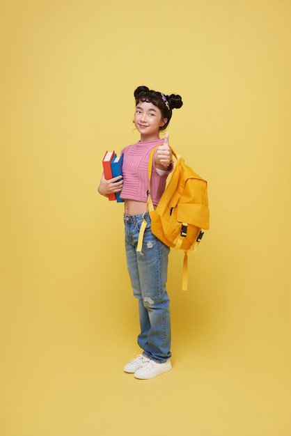 Portrait of happy smiling young Asian student elementary school girl with notebook and backpack ready to school isolated on yellow background