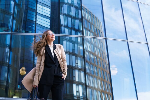 Portrait of a happy successful caucasian ethnicity woman in glasses stands in a coat and suit agains