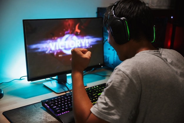 Portrait of happy teenage gamer boy winning while playing video games on computer in dark room, wearing headphones and using backlit colorful keyboard