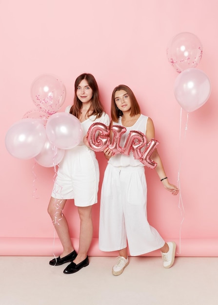 Portrait of happy two young women dressed in a white holdings a balloons over pink background