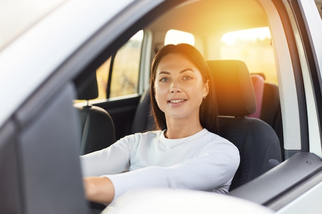 Portrait of happy woman driving a car and smiling cute young success brunette woman driver steering car wearing white shirt looking at camera with satisfied expression