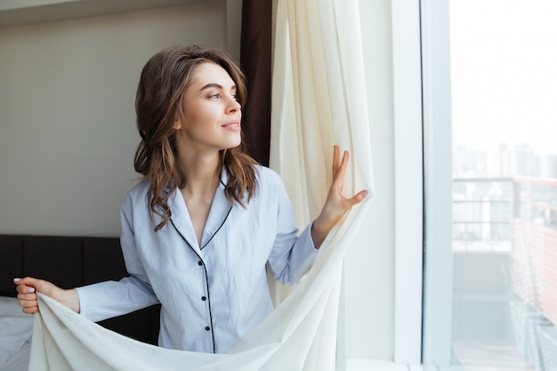 Portrait of a happy woman opening window curtains