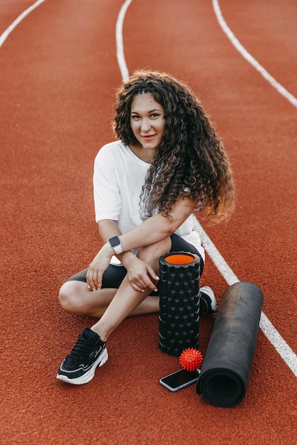 Portrait of a happy woman sitting on a running track near yoga mat, mfr roller, her phone, mfr ball in the morning.