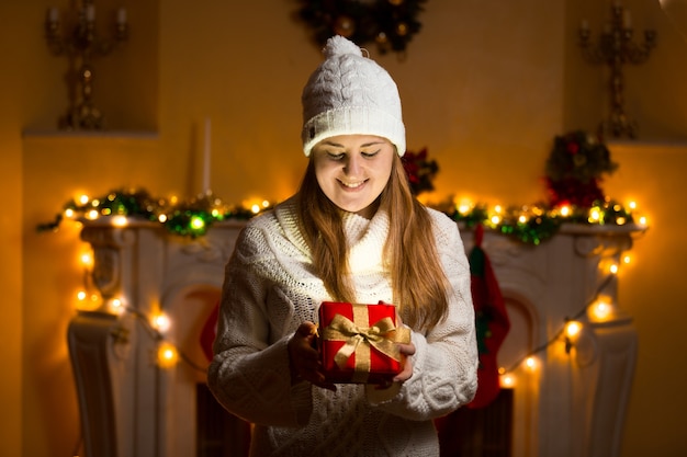 Portrait of happy woman in sweater holding glowing gift box at Christmas eve