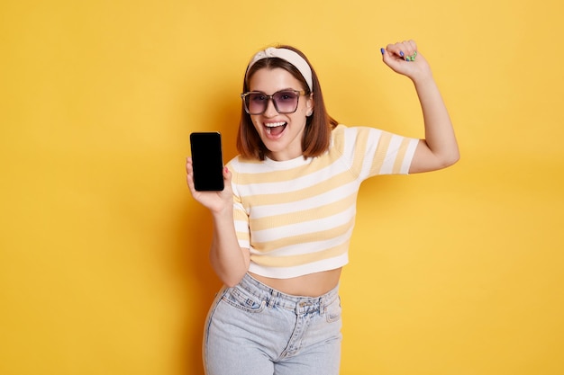 Portrait of happy woman wearing striped t shirt and jeans posing isolated over yellow background showing blank black screen of smart phone and clenched fist copy space for advertisement