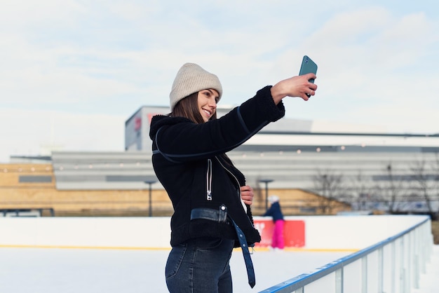 Portrait of a happy woman in winter cloth making selfie photo on smartphone while skating on the ice