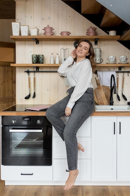 Portrait of happy woman with laptop sitting on modern light wood kitchen furniture in casual clothes