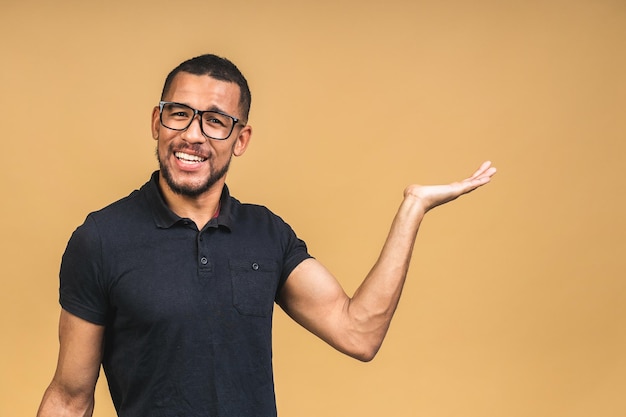 Portrait of happy young african american black man with in casual smiling pointing aside with finger looking in camera with excited face expression isolated over beige background