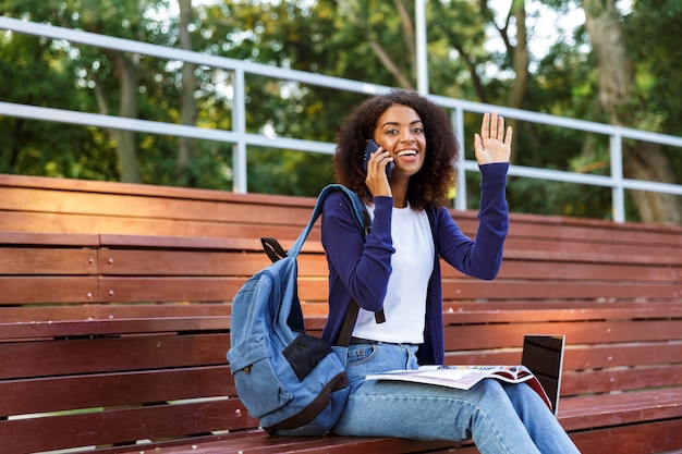 Portrait of a happy young african girl with backpack talking on mobile phone while resting at the park, reading magazine, waving hand