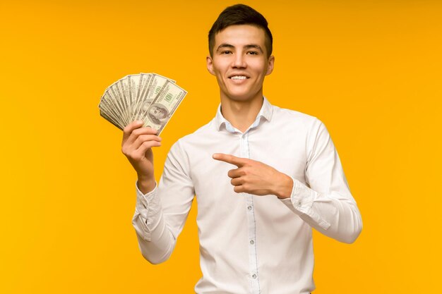 Portrait of happy young asian man holding credit card and money in hand smiling and looking at camera on isolated yellow background feeling positive and enjoy image