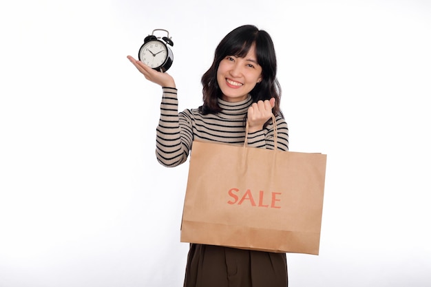 Portrait of happy young Asian woman with sweater shirt holding alarm clock and shopping paper back isolated on white background