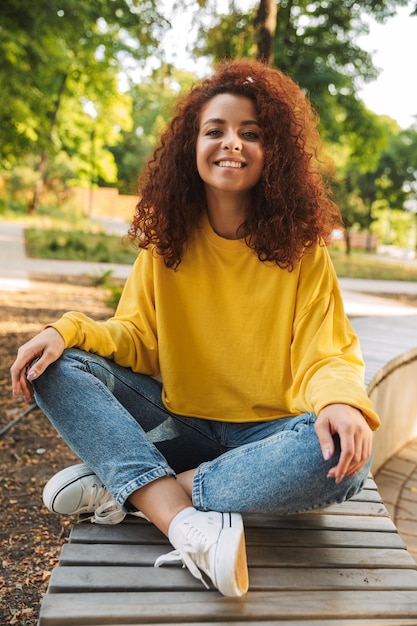 Portrait of a happy young beautiful curly student outdoors in nature park sitting on a bench.