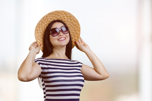 Portrait of happy young brunette woman wearing sunglasses and straw hat bright blurred background