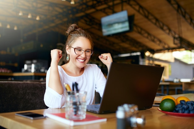 Portrait of happy young business woman celebrating success with arms up in front of laptop