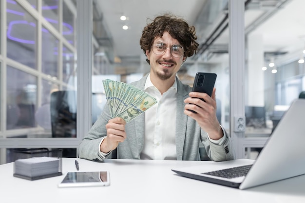 Portrait of happy young businessman man sitting at desk in office holding and showing phone and