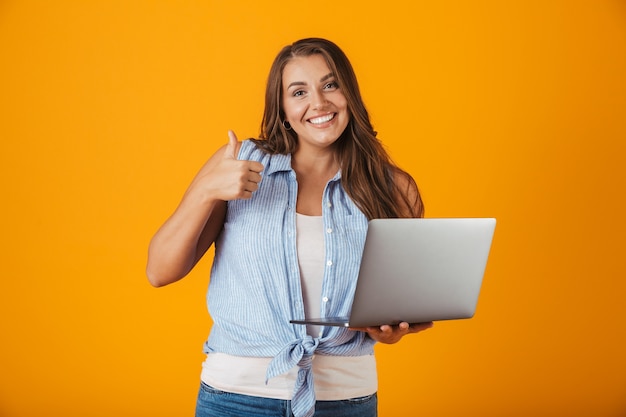 Portrait of a happy young casual woman, holding laptop computer, giving thumbs up