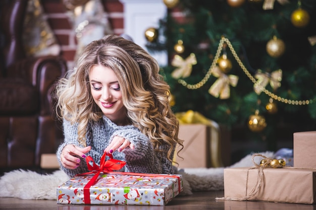 Portrait of a happy young Christmas woman trying to guess what is in the gift box near the Christmas tree lying on the floor in the living room Close it