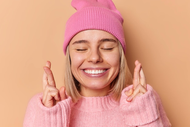 Portrait of happy young European woman closes eyes smiles broadly shows white teeth keeps fingers crossed believes in good luck has cherishes wish hopes for forune isolated over brown background