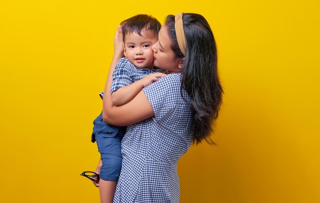 Portrait of happy young family mother hugging and kissing son child isolated on yellow background Mother's Day parenthood childhood concep