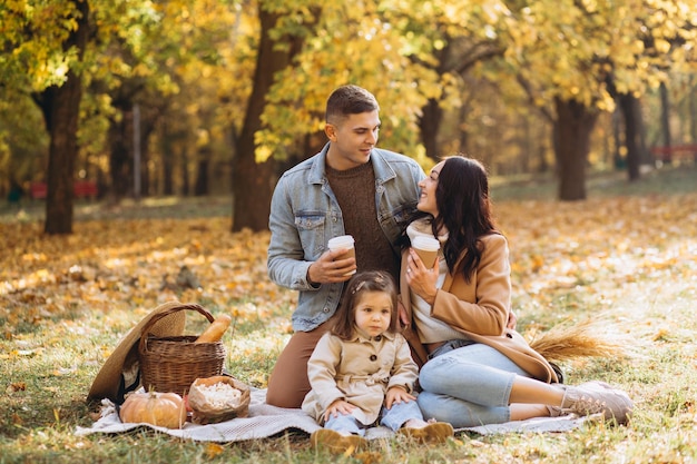 Portrait of happy young family sitting on plaid together and drinking cacao in autumn park