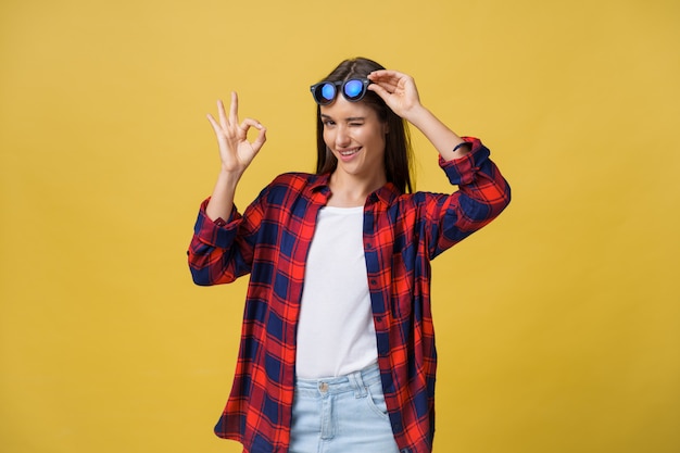 Portrait of a happy young girl in summer clothes showing ok gesture over yellow background.