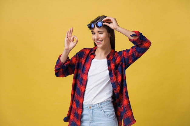 Portrait of a happy young girl in summer clothes showing ok gesture over yellow background.