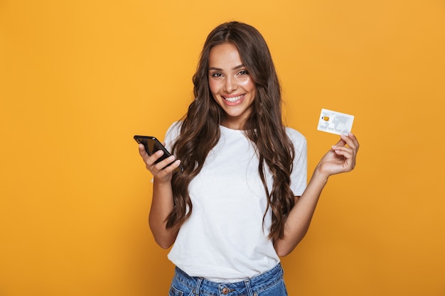 Portrait of a happy young girl with long brunette hair standing over yellow wall, holding mobile phone, showing plastic credit card