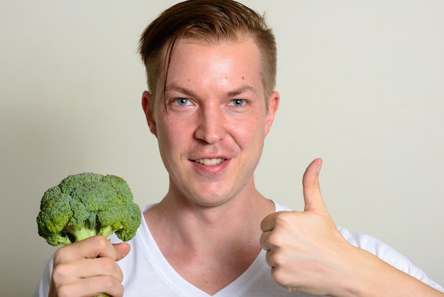 Portrait of happy young handsome man holding broccoli and giving thumbs up