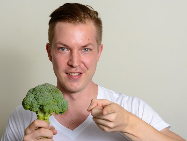 Portrait of happy young handsome man holding broccoli and pointing at camera