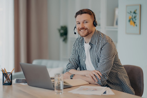 Portrait of happy young male freelancer sitting at his modern cozy workplace and working on laptop, cheerful guy smiling at camera and enjoying remote job. Freelance concept