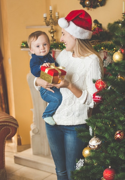 Portrait of happy young mother cuddling her baby boy in Santa cap at Christmas tree