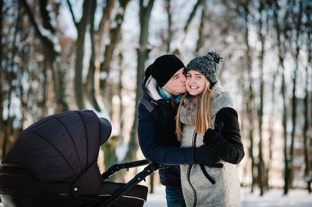 Portrait of a happy young parents stand and kissing with a stroller baby in a winter park