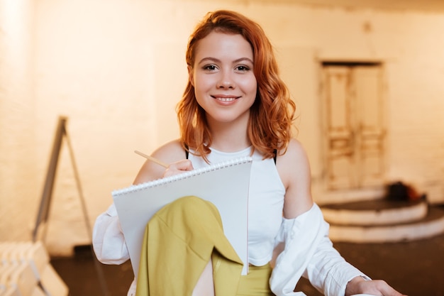 Portrait of happy young redhead lady painter with album in artist workshop.