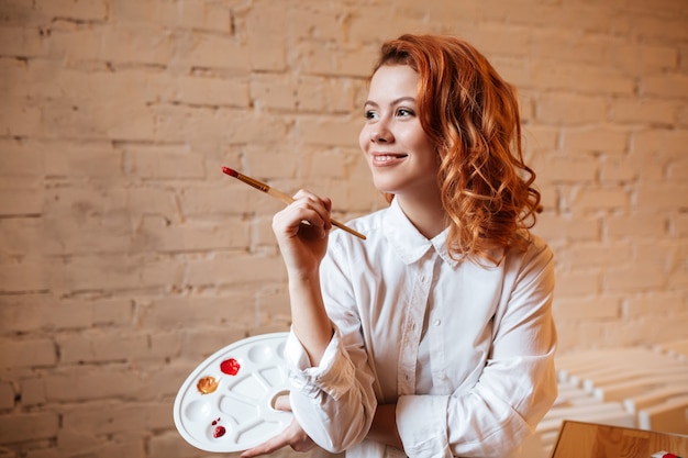 Portrait of happy young redhead woman painter with oil paints and palette.