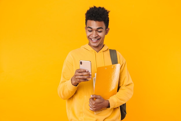 Portrait of a happy young teenager boy standing isolated over yellow wall, carrying backpack and textbooks, using mobile phone